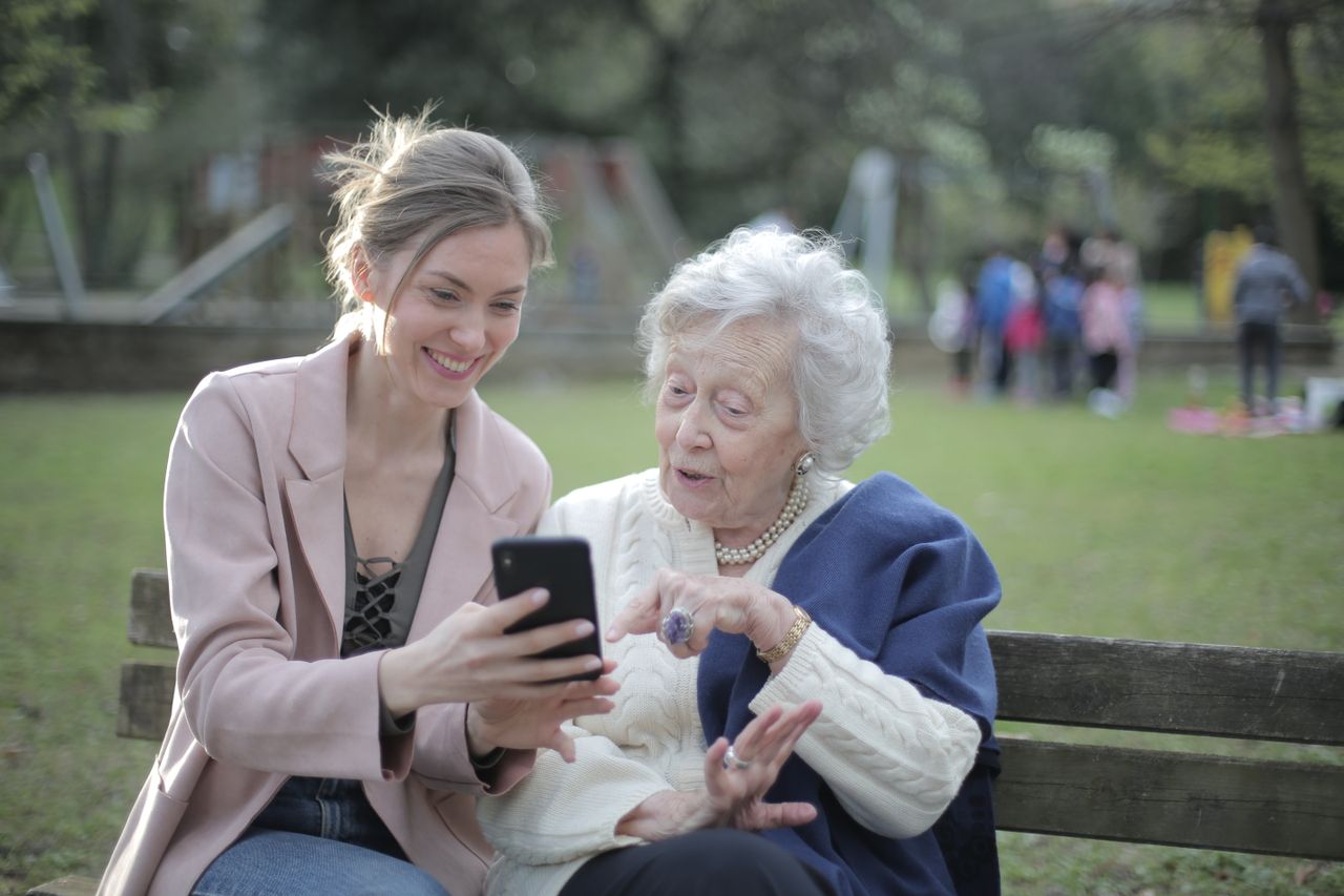 An older woman and her adult daughter sitting on a park bench, smiling at a phone together
